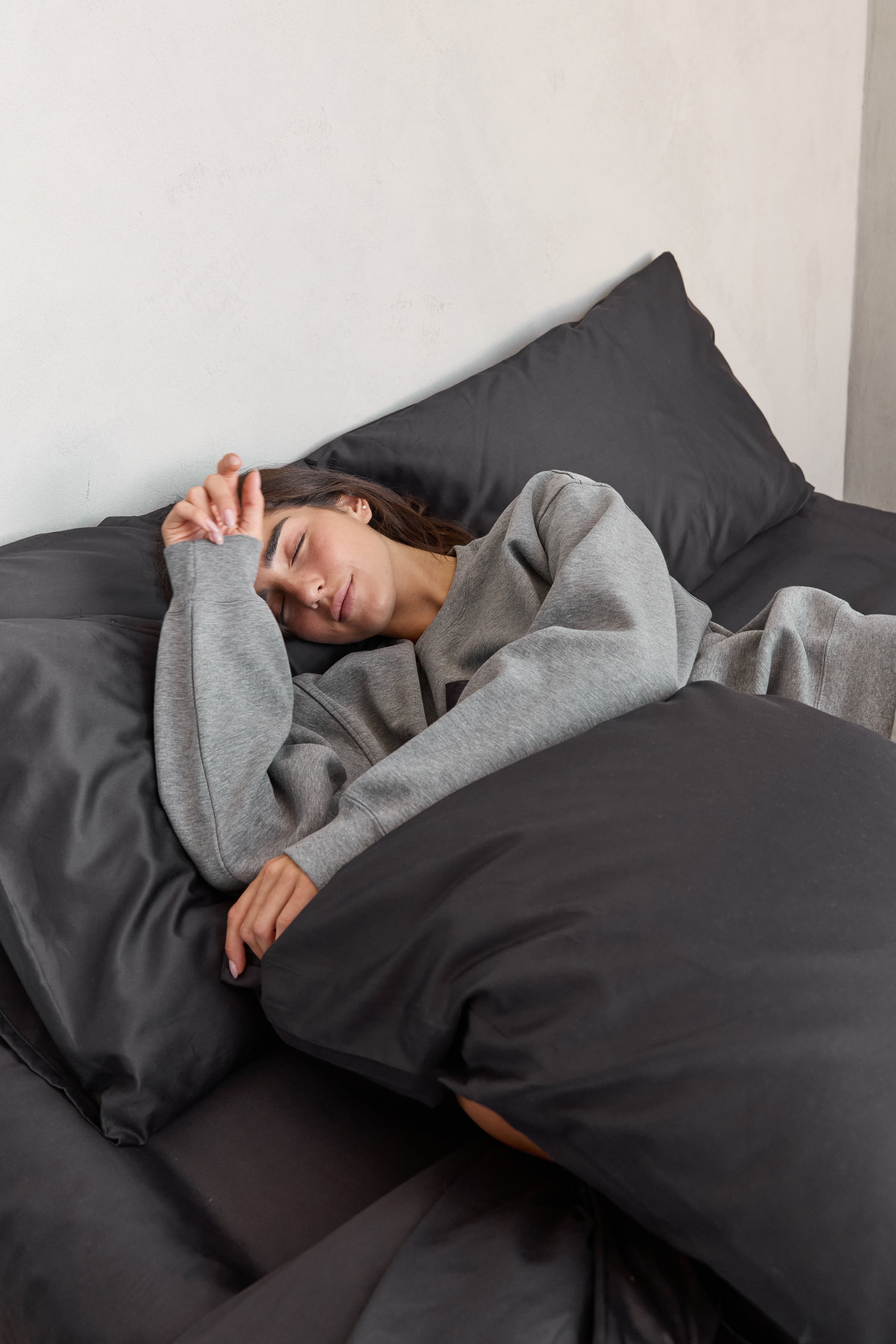 דוגמנית שוכבת על מיטה עם מצעי כותנה בצבע אפור פחם ומחבקת כרית