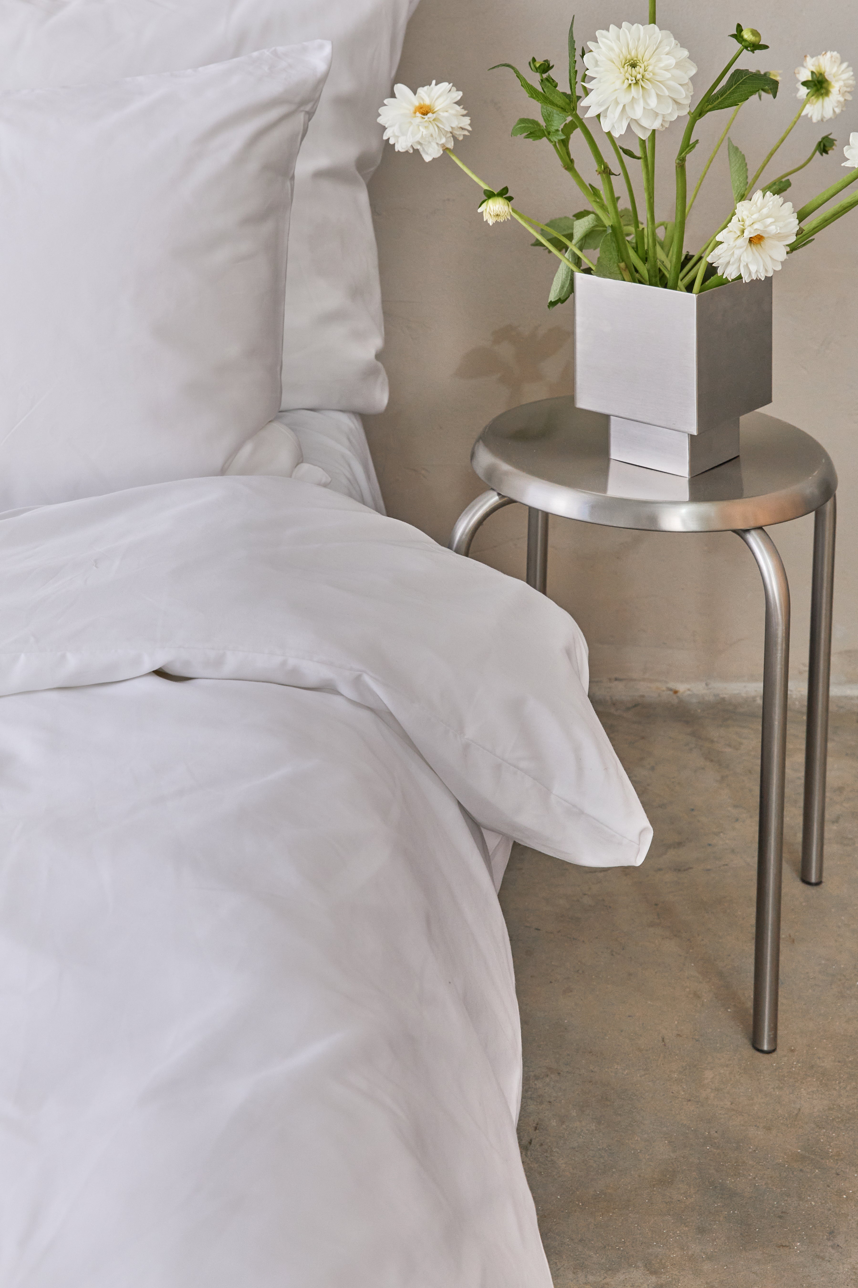 תקריב צד ימין של מיטה זוגית עם מצעי כותנה לבנים, שרפרף מתכת ועליו ואזה ממתכת עם זר פרחים לבנים