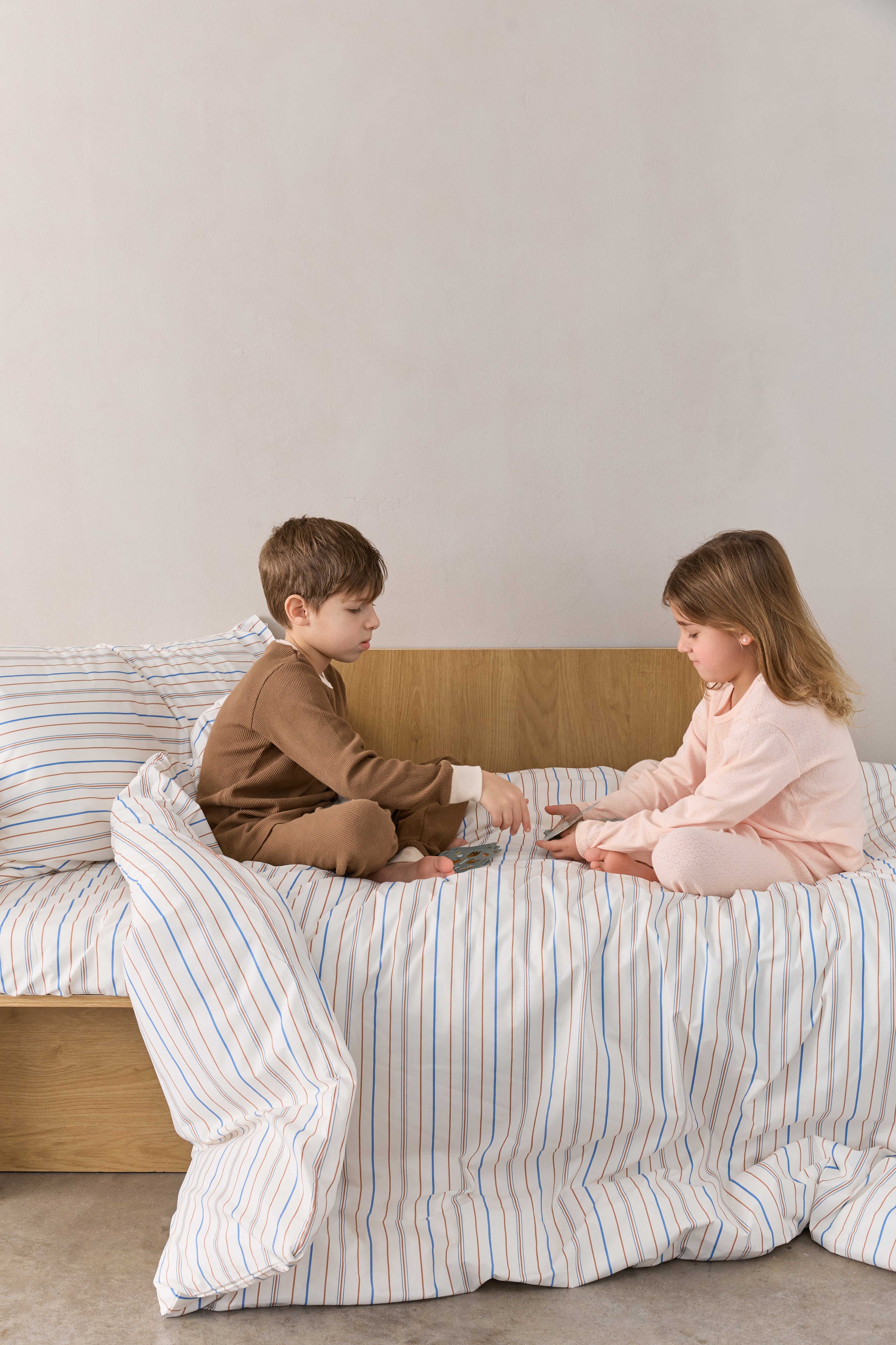 ילד וילדה יושבים על מיטה ומשחקים בקלפים, על המיטה סט מצעי פרקל בצבע קרם עם פסים חומים וכחולים