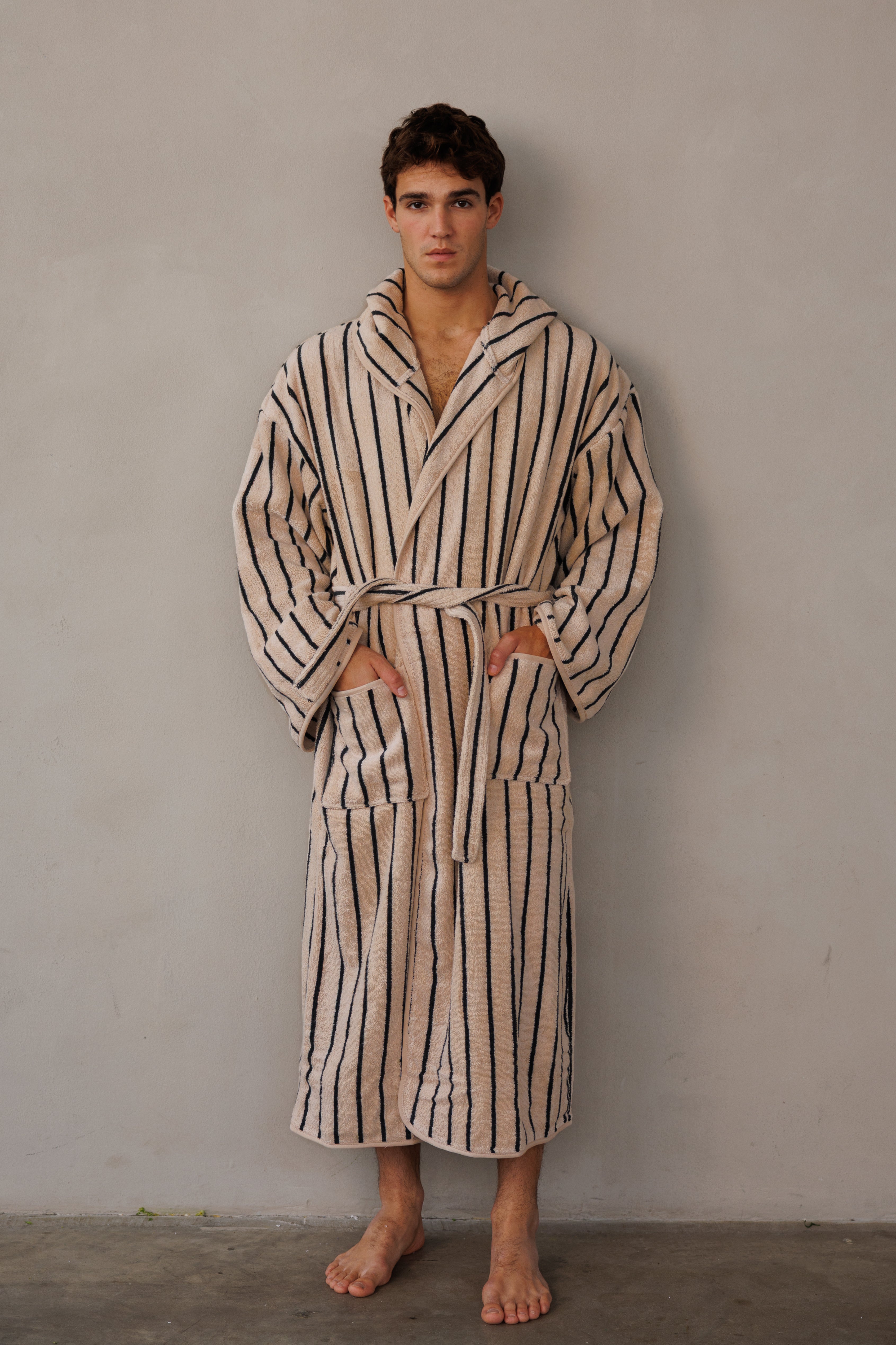 דוגמן עומד כנגד קיר עם חלוק בצבע בז' עם פסים שחורים וידיים בכיסים