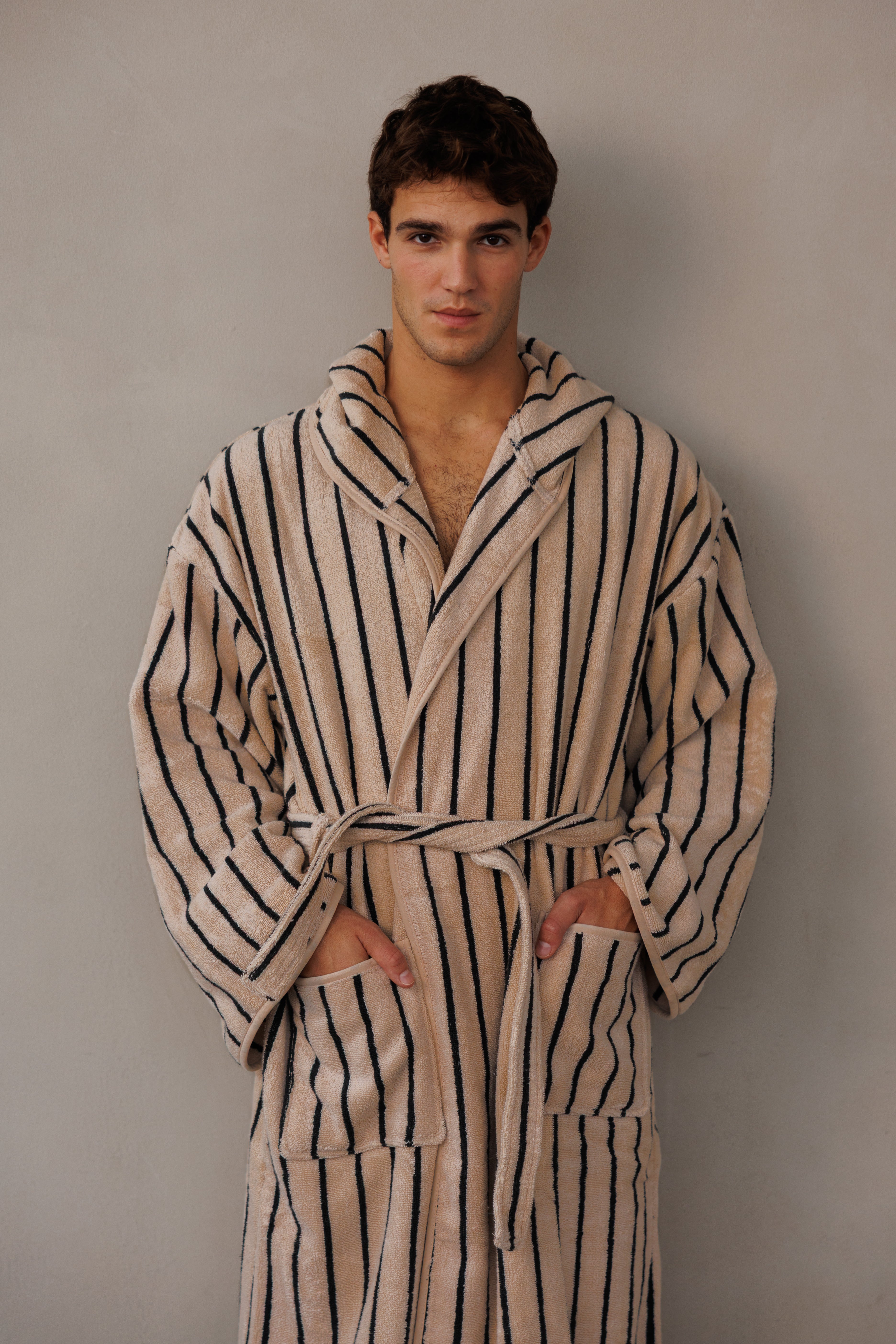 תקריב דוגמן עומד כנגד קיר עם חלוק בצבע בז' עם פסים שחורים וידיים בכיסים