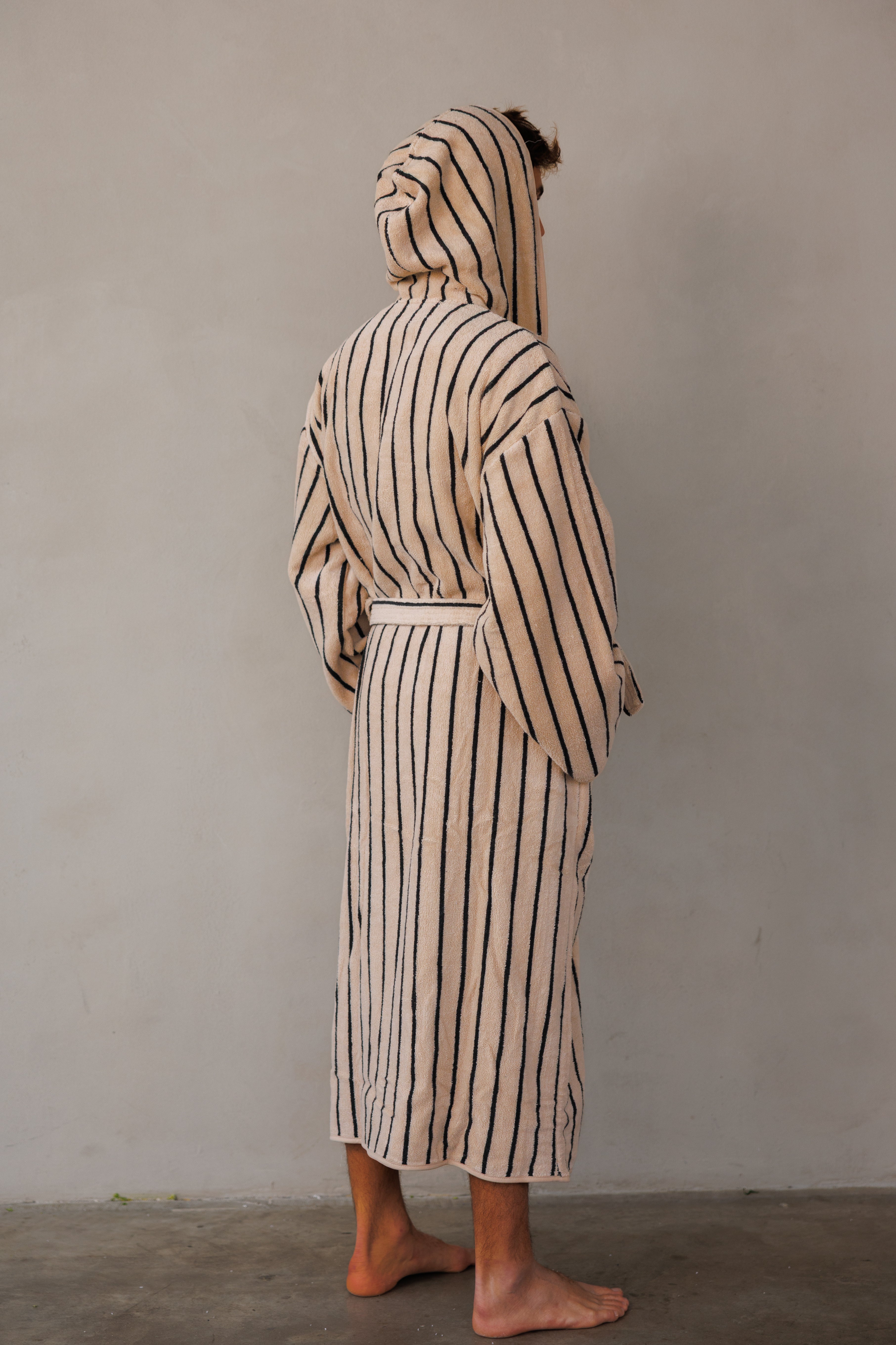 דוגמן עומד עם גב למצלמה עם חלוק בצבע בז' עם פסים שחורים ,ידיים בכיסים וכובע החלוק על הראש