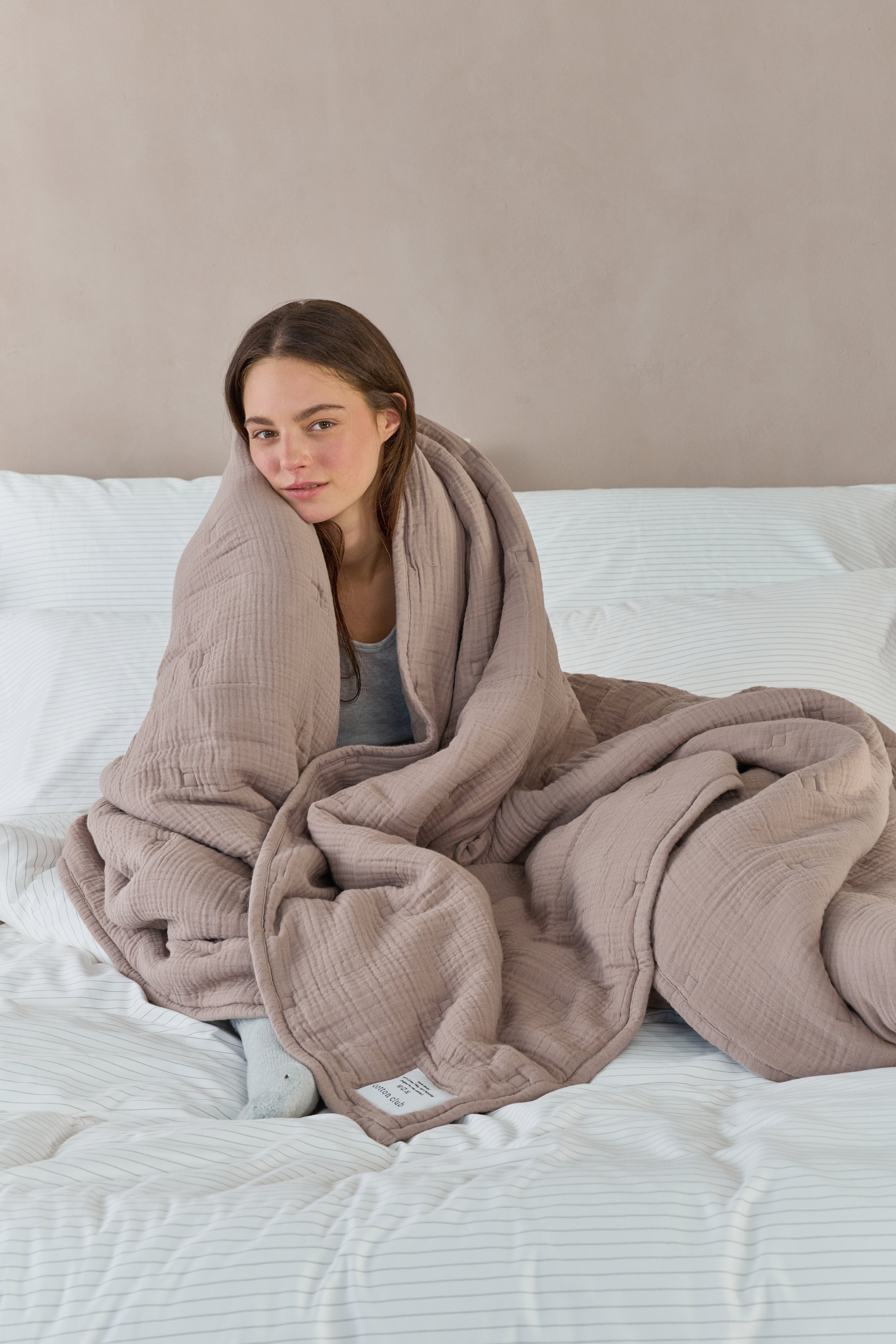 דוגמנית יושבת על מיטה עם מצעים לבנים עטופה בשמיכת פוך מוסלין בצבע מוקה