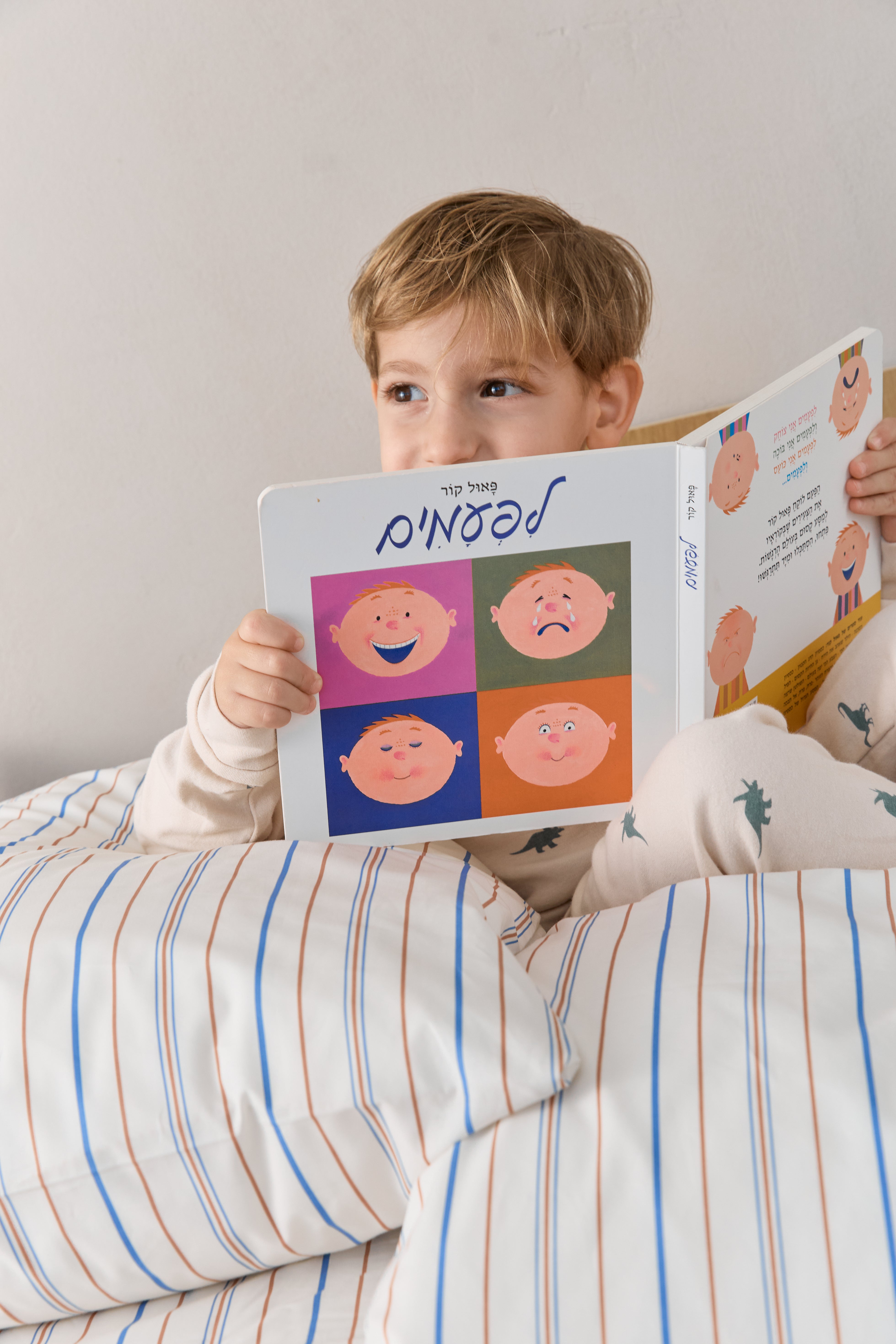 ילד יושב על המיטה וקורא ספר, על המיטה סט מצעי פרקל בצבע קרם עם פסים חומים וכחולים