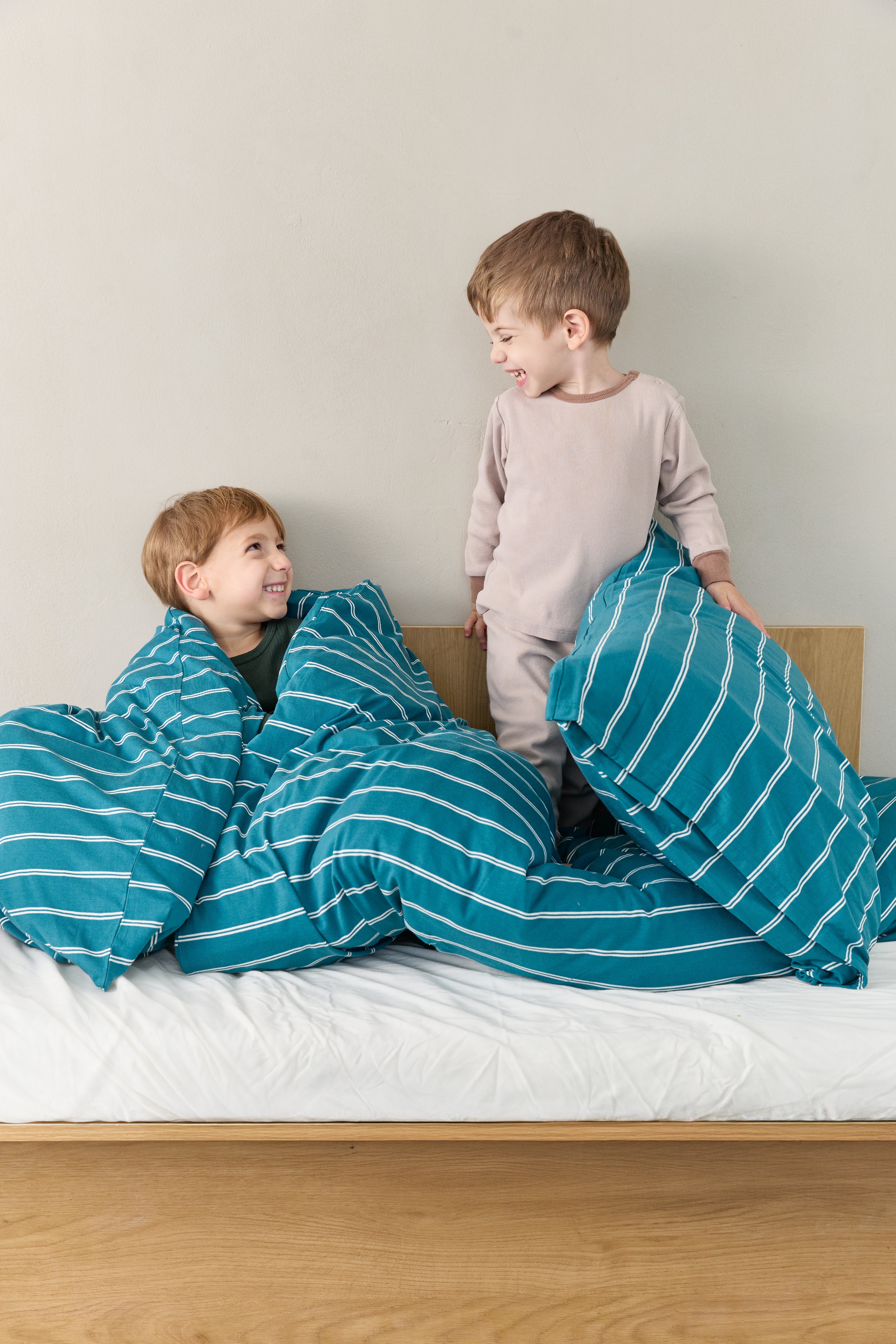 שני ילדים יושבים על מיטת ילדים מסתכלים אחד על השני ומחייכים, על המיטה מצעי ג'רסי במבוק בצבע טורקיז עם פסים לבנים, הסדין לבן חלק