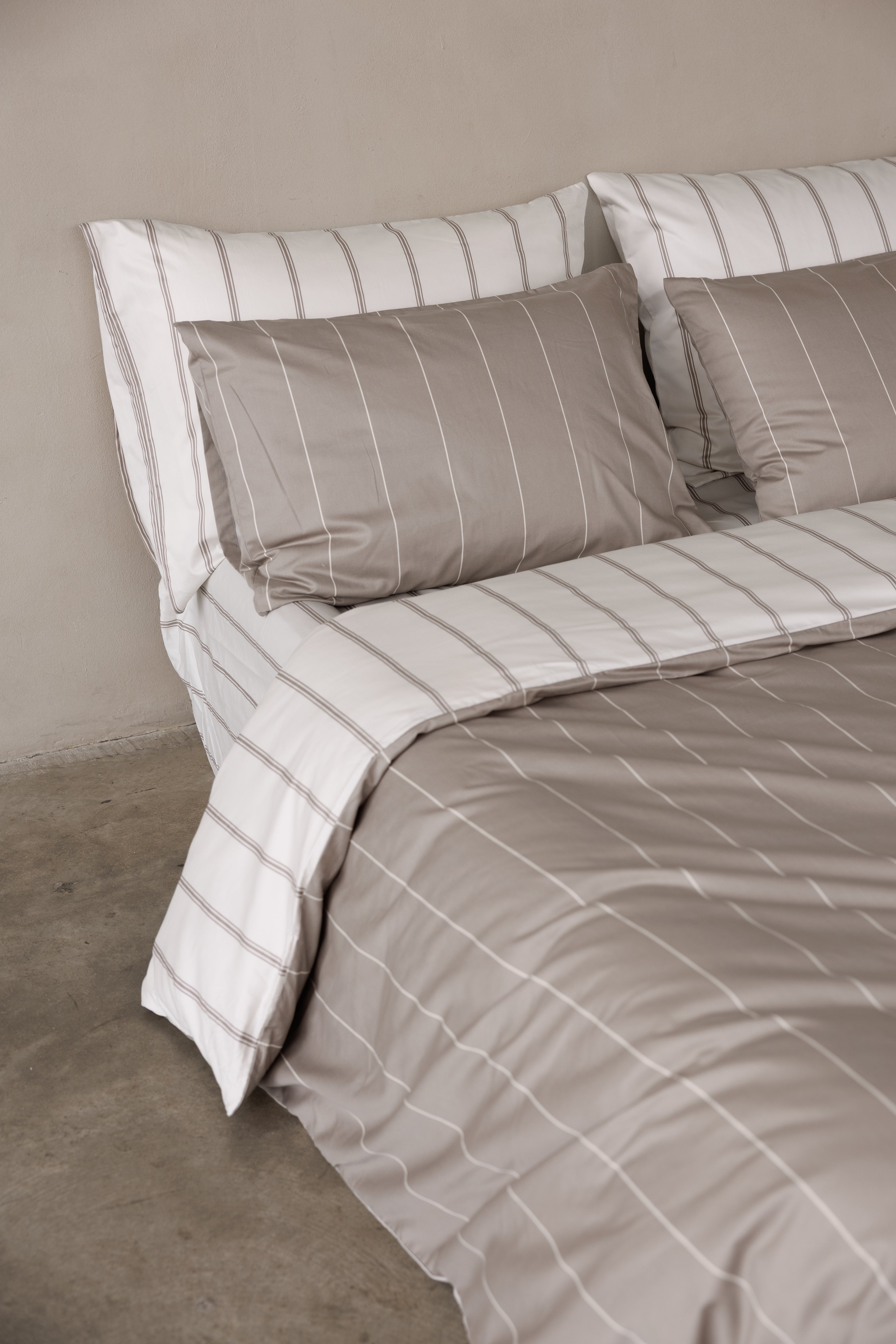 פינה של מיטה זוגית עם מצעי סאטן 500, כריות אחוריות בצבע קרם עם פסים אבן וכריות קדמיות בצבע אבן עם פסים קרם. הציפה דו צדדית צד אחד אבן עם פסים קרם וצד שני קרם עם פסים אבן