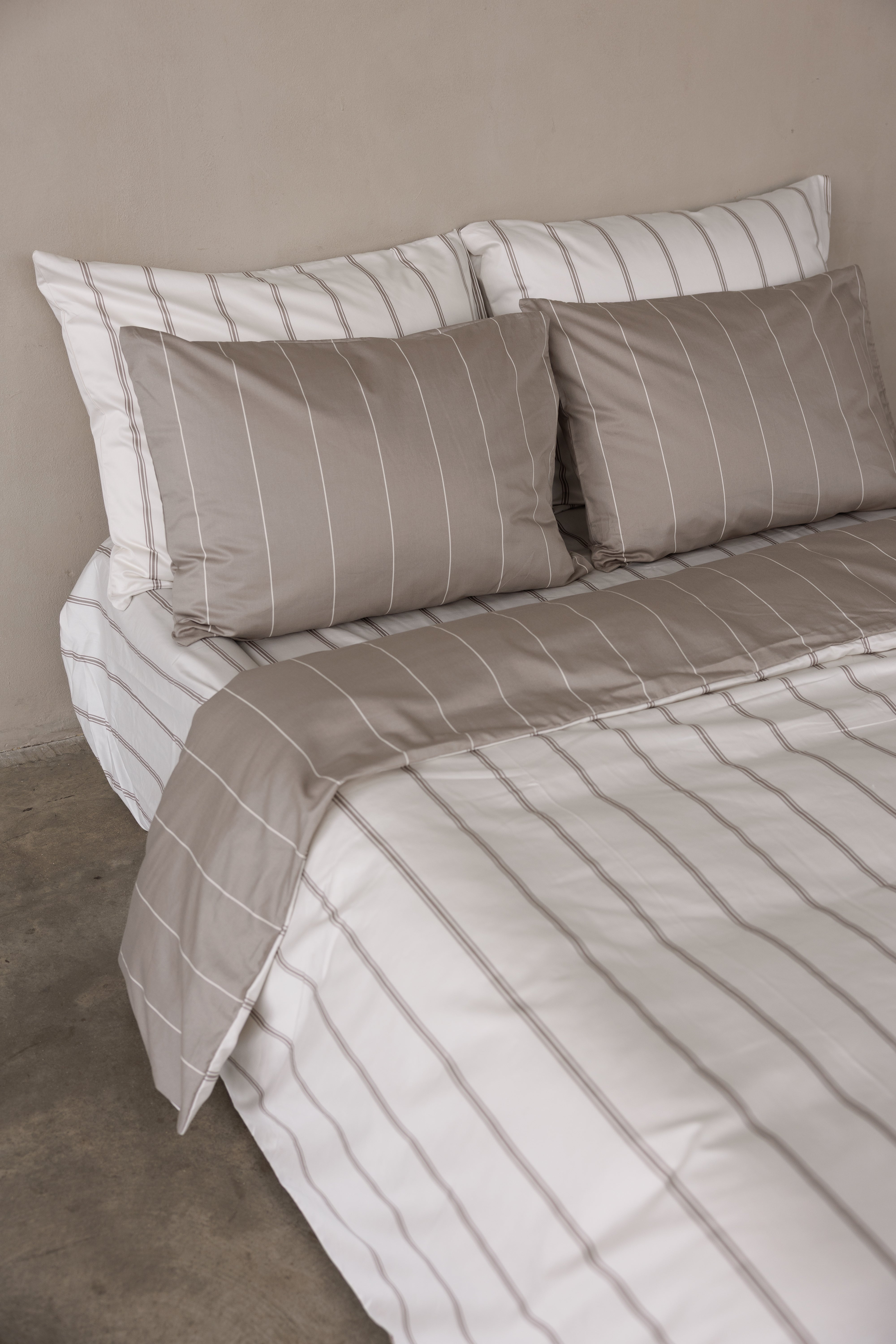 פינה של מיטה זוגית עם מצעי סאטן 500, כריות אחוריות לבנות עם פסים בצבע אבן , כריות קדמיות בצבע אבן עם פסים לבנים. הציפה דו צדדית צד אחד אבן עם פסים לבנים וצד אחד לבן עם פסים אבן