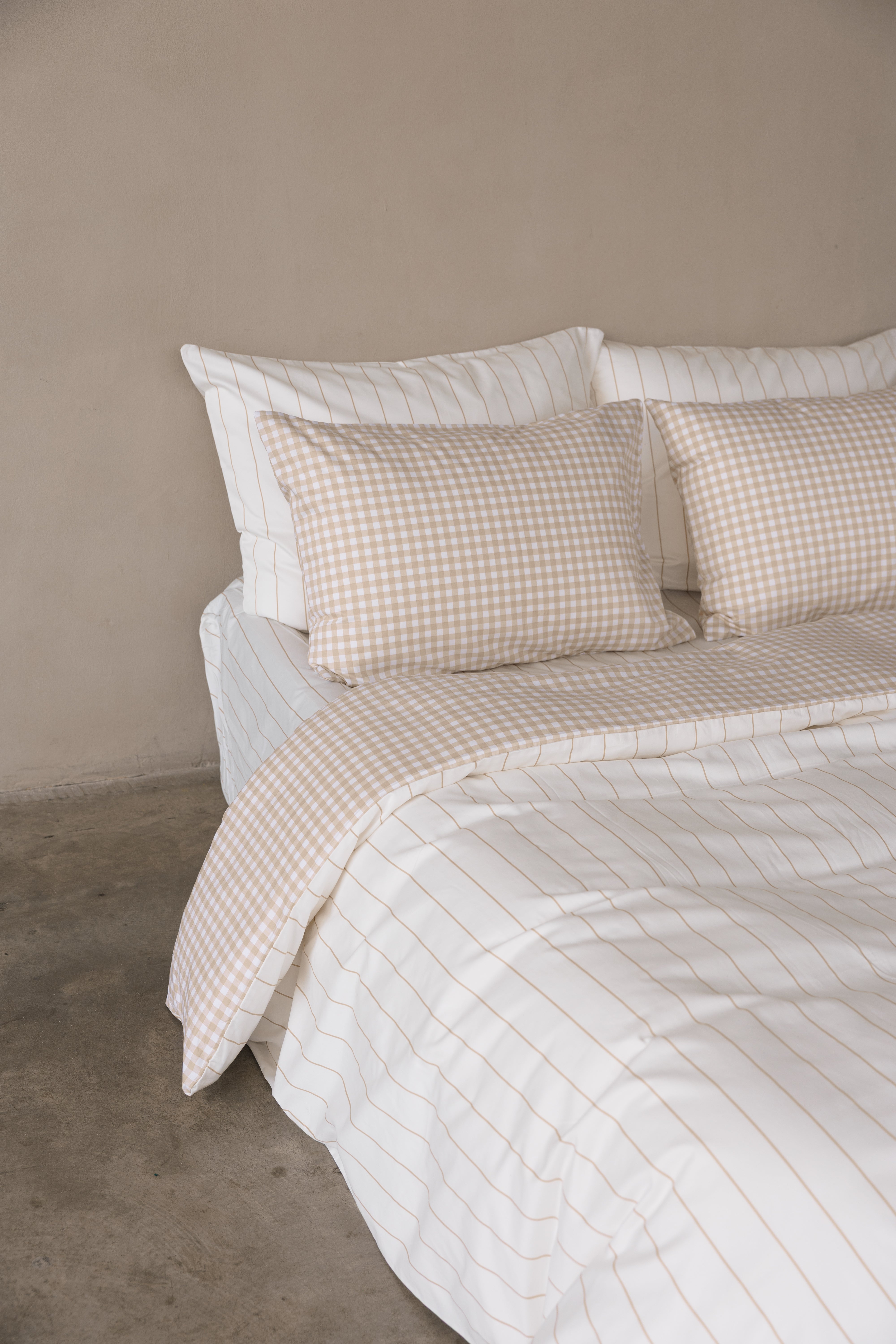 פינה של מיטה זוגית עם מצעי סאטן 500 כריות אחוריות לבנות עם פסים בז', כריות קדמיות משבצות בז', סדין לבן עם פסים בז' וציפה דו צדדית צד אחד משבצות צד אחד פסים